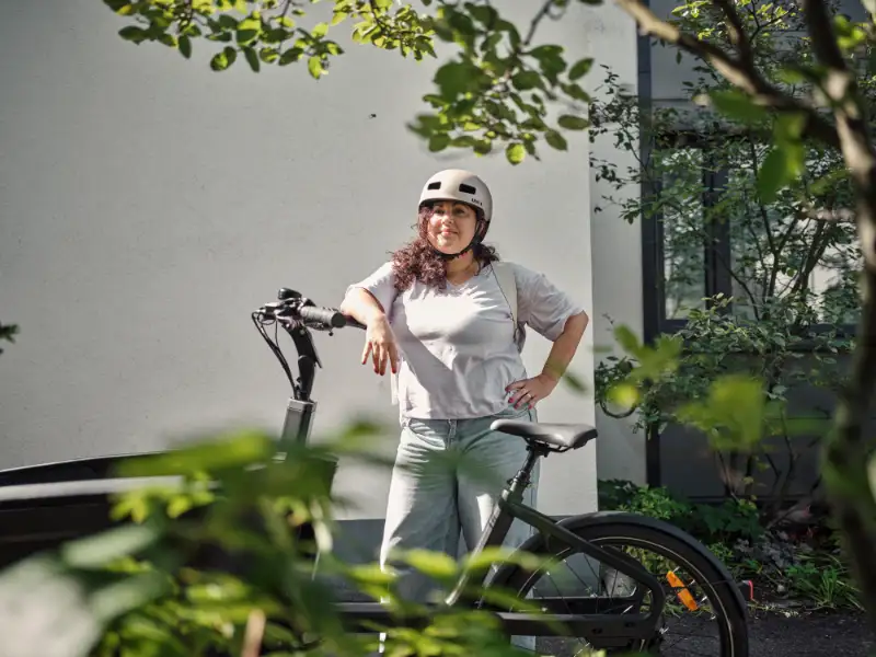 Eine Frau lehnt stolz an ihrem Lastenrad und trägt einen Helm.