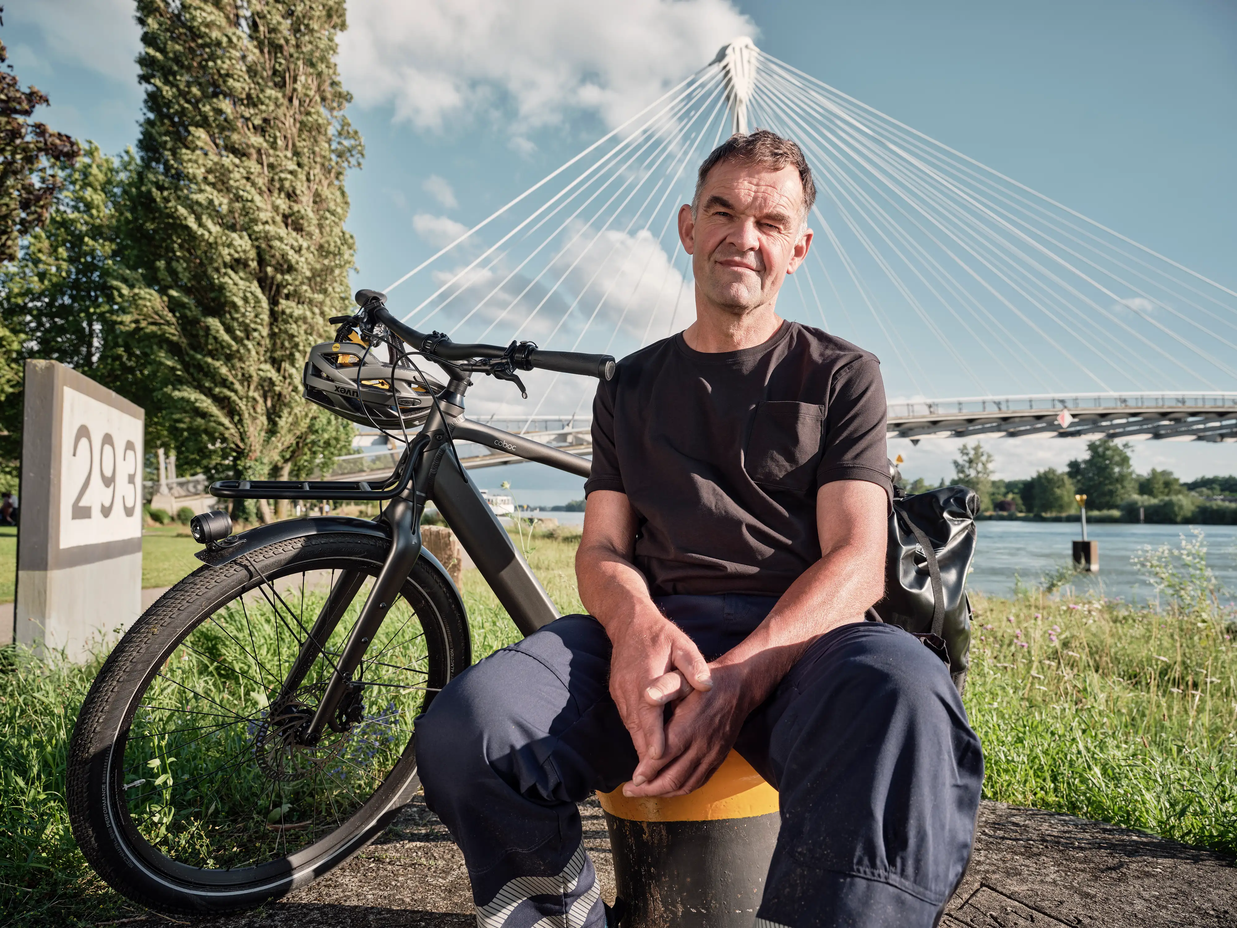 Ein Mann sitzt vor einer Brücke auf einem Poller. Hinter ihm steht sein Fahrrad.